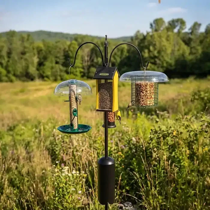 Three bird feeders on a stand in a natural setting with trees and grass.